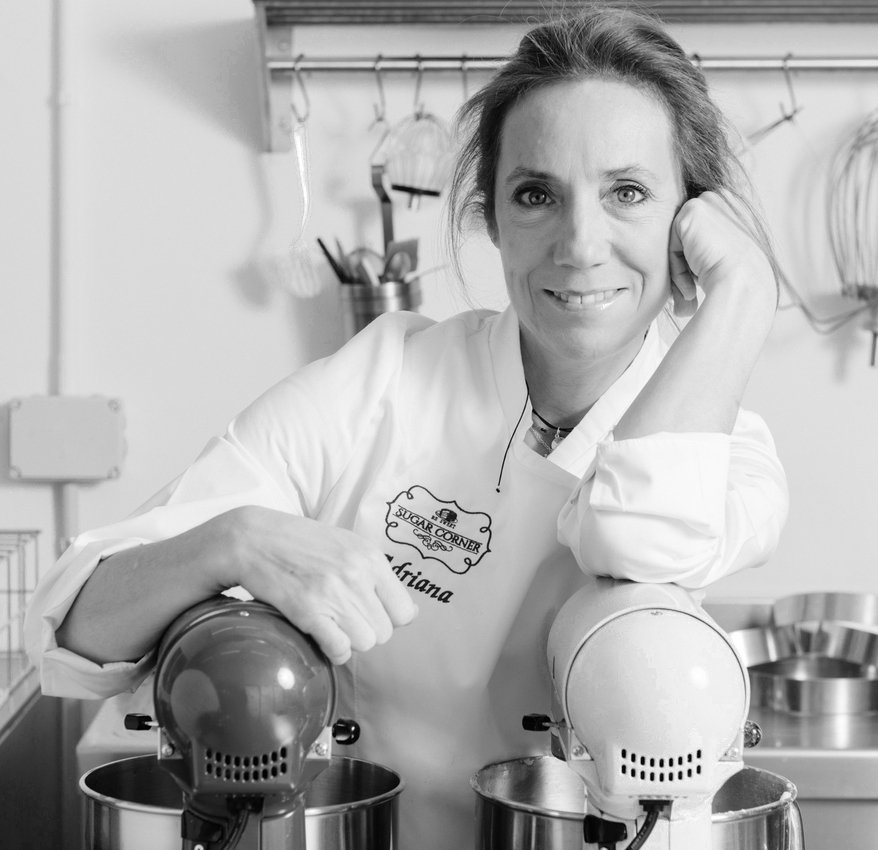 Una mujer con una bata de chef blanca se apoya en una batidora de pie roja y azul en una cocina con utensilios de acero inoxidable en la pared detrás de ella.