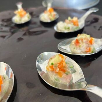 Varias cucharas de metal contienen cada una una pequeña porción de una mezcla de verduras y pescado finamente picados, dispuestos sobre una superficie oscura.