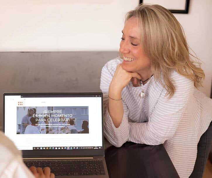 Una mujer se sienta en una mesa, sonriendo y mirando la pantalla de una computadora portátil que muestra un sitio web de pastelería creativa con el texto “SIEMPRE ES BUEN MOMENTO PARA CELEBRAR”.