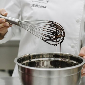 Una persona con una bata de cocinero blanca sostiene un batidor cubierto de chocolate encima de un recipiente de metal lleno de chocolate derretido.