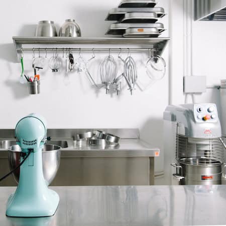 Una cocina comercial con una batidora de pie azul en primer plano, utensilios de metal y accesorios para mezclar colgados en una rejilla y una gran batidora industrial a la derecha, perfecta para la formación de equipos en Barcelona o para la preparación de experiencias gourmet.