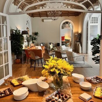 Un acogedor salón con asientos modernos, grandes ventanas arqueadas, techo ornamentado y una mesa de madera con bocadillos, flores y cuencos en primer plano.