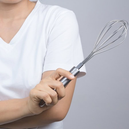 Persona con una camisa blanca sosteniendo un batidor de metal en su mano derecha sobre un fondo gris liso, perfecto para un evento de pastelería creativa o de team building en Barcelona.