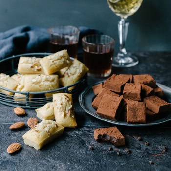 Una cesta de alambre con pasteles de almendras y un plato de trufas de chocolate espolvoreadas con cacao sobre una superficie oscura, con bebidas de fondo.
