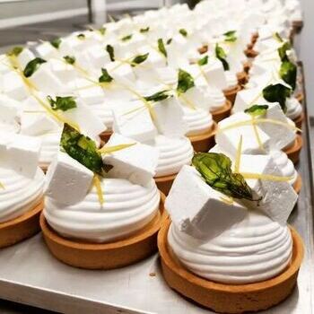 Filas de tartas redondas de hojaldre cubiertas con crema blanca, grandes cubos blancos, hojas verdes y finas tiras amarillas muestran la pastelería creativa, perfecta para una experiencia gastronómica gourmet o su próximo evento de formación de equipos en Barcelona.