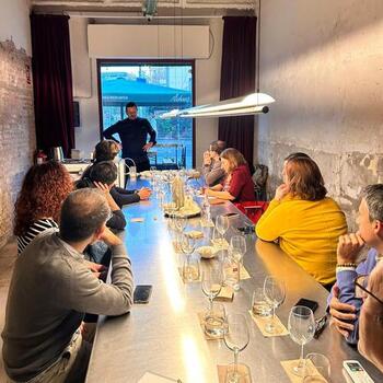 Un grupo de personas se sienta alrededor de una mesa larga con copas de vino vacías, escuchando a una persona que habla al frente de la sala.