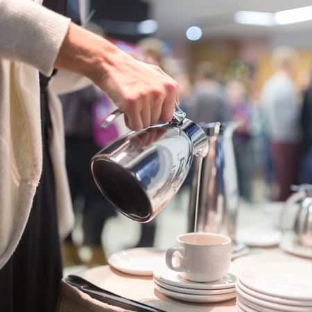 Una persona vierte café de una cafetera de metal en una taza blanca en una mesa preparada con platos y tazas, evocando el ambiente de un restaurante gourmet, con gente borrosa en el fondo.