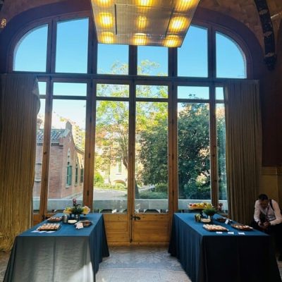 Frente a grandes ventanales arqueados con vistas al exterior se disponen dos mesas con refrescos gourmet, detrás de una de las mesas hay una persona.