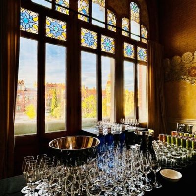Una mesa preparada con copas de vino vacías y un gran cuenco de metal se encuentra frente a altos vitrales, ofreciendo el entorno perfecto para realizar actividades de team building en Barcelona o explorar la gastronomía gourmet con vistas a un encantador patio.