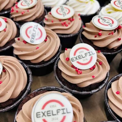 Cupcakes de chocolate con glaseado marrón claro, chispas rojas y adornos redondos con la etiqueta "EXELFIL 2024" dispuestos muy juntos: perfectos para catering de eventos gourmet o actividades de team building en Barcelona.