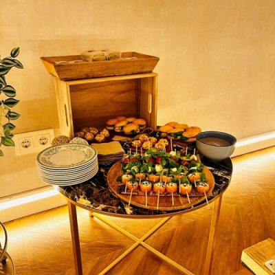 Una mesa redonda muestra aperitivos gourmet en brochetas, pasteles inspirados en la pastelería creativa, platos apilados, cuencos y una caja de madera, todo ello contra una pared beige con una iluminación suave.