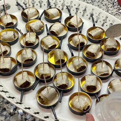 Una bandeja con varias cucharitas, cada una con un pequeño aperitivo envuelto en una cobertura blanca, servido en un líquido amarillo con un palillo. Ideal para catering de eventos gourmet o actividades de team building en Barcelona. Cerca, una mano sostiene un recipiente de plástico.