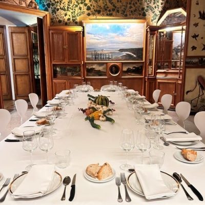 Una mesa de comedor formal preparada para una comida con mantel blanco, cristalería, cubiertos y pan; el centro está compuesto por calabazas y verduras. El salón, decorado con paneles de madera y arte mural, es perfecto para una actividad de team building en Barcelona o para un evento gastronómico con catering.