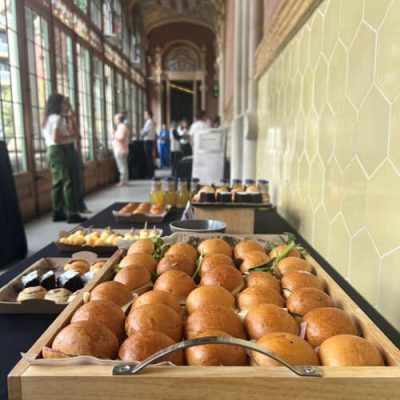 Bandejas de panecillos variados y pasteles de pastelería creativa en una mesa de buffet en un gran salón, con gente mezclándose en el fondo durante un evento de Team Building en Barcelona.