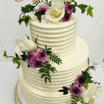 Pastel de dos niveles con glaseado blanco decorado con rosas blancas, flores moradas, hojas verdes y un adorno que dice "amor": una elegante creación de nuestra pastelería creativa, perfecta para catering de eventos gourmet o celebraciones especiales.