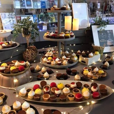 Una mesa de postres muestra una variedad de coloridos mini pasteles y tartas de pastelería creativa en bandejas escalonadas, rodeadas de plantas, decoración y luces de cadena en un ambiente interior moderno.