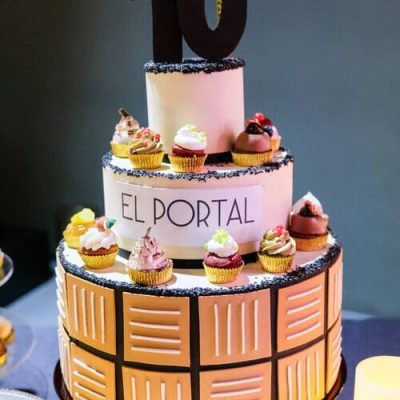 Una tarta de dos pisos de nuestra pastelería creativa, con el "10" en la parte superior, cupcakes como decoración y "EL PORTAL" en el piso central. El piso inferior presenta un diseño geométrico, perfecto para catering de eventos gourmet o de team building en Barcelona.