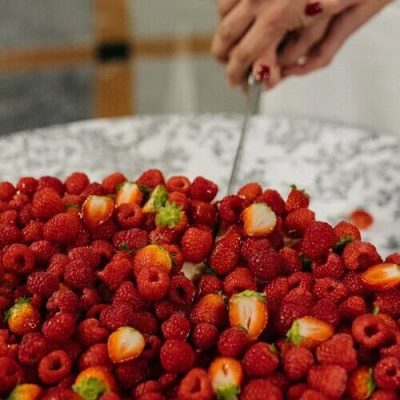 Dos personas cortando un pastel cubierto de frambuesas y fresas frescas sobre un mantel blanco, mostrando el arte de la pastelería creativa en la alta gastronomía gourmet.