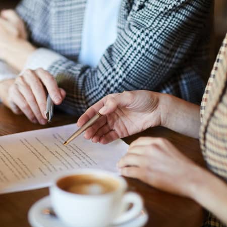 Dos personas se sientan en una mesa revisando un documento juntas, cada una sosteniendo un bolígrafo, con una taza de café en primer plano: perfecto para una sesión de formación de equipos en Barcelona o para discutir opciones gourmet de catering.